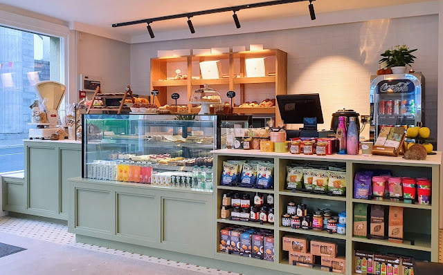 Cafe counter with weighing scales, a glass-front display area for chilled cakes and sandwiches, scones and muffins displayed on shelves being the counter, and a retro-style coke fridge for cold drinks.  Gourmet jams and spreads, packets of crisps and oatmeal cookies on the display shelving