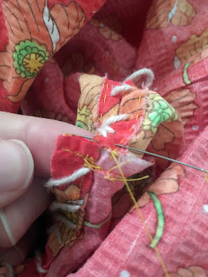 A close-up of a needle inserted into the seam allowance of a coral piece of fabric with scattered orange poppy motifs, with a white finger holding up the stitched-shut end of the seam allowance. A white, slightly fuzzy cord is laced diagonally through the seam allowance, attaching it to another finished edge.