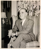 A photograph of an older Flanigan sitting in a chair looking bemused