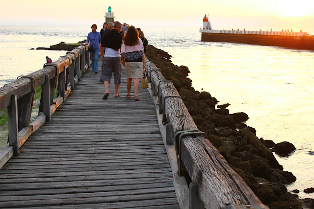 letnim wieczorem na drewnianym molo w Capbreton, spacerujący po molo turyści, w tle dwie latarnie znaczące wejście do portu, niebo zabarwione lekko czerwowanym światłem zachodzącego słońca 