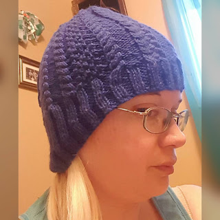 Close up of a side view; a blonde woman wears a blue hat with graduated ribbing (longer in the back than the front) and a pattern of cables and moss stitch which narrow towards the top