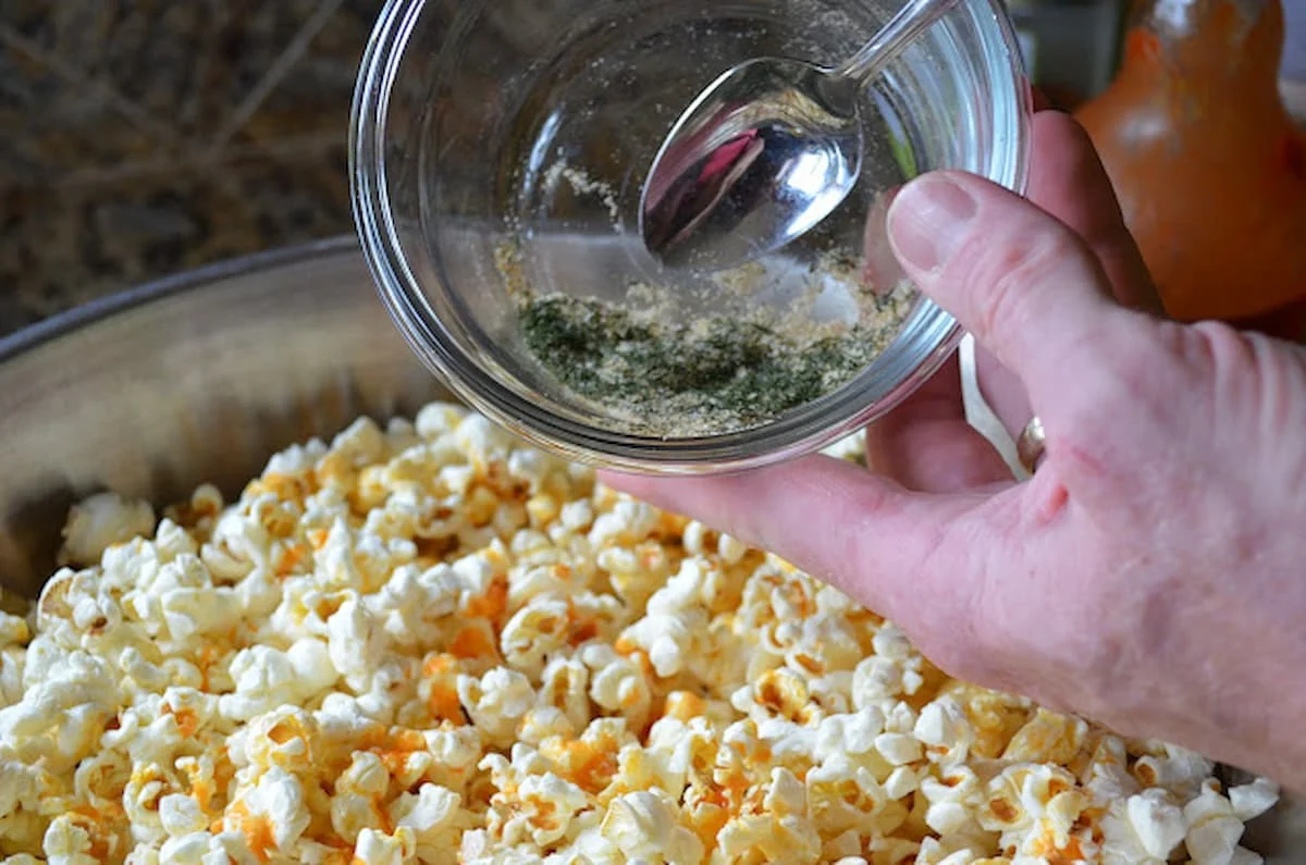 A stainless steel bowl of Buffalo Ranch Popcorn with ranch seasoning being sprinkled over the top.