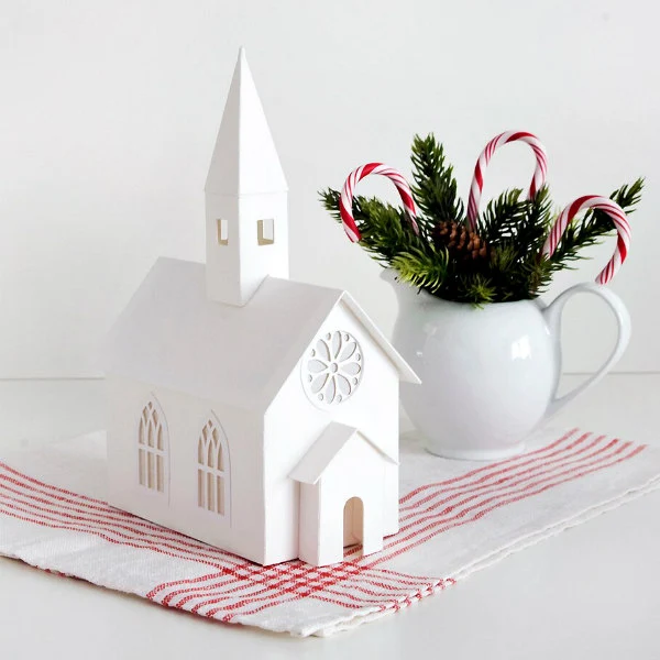 miniature white card stock rose window putz church displayed on red and white tea towel and placed next to white pitcher full of greenery and candy canes.