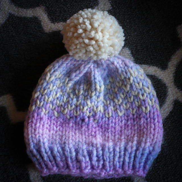 Flat shot of a blue and lavender gradation knit hat with a cream pom pom and cream hearts on a gray and white rug.