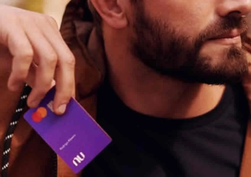 Imagem mostra um homem segurando o cartão do nubank entre seus dedos.