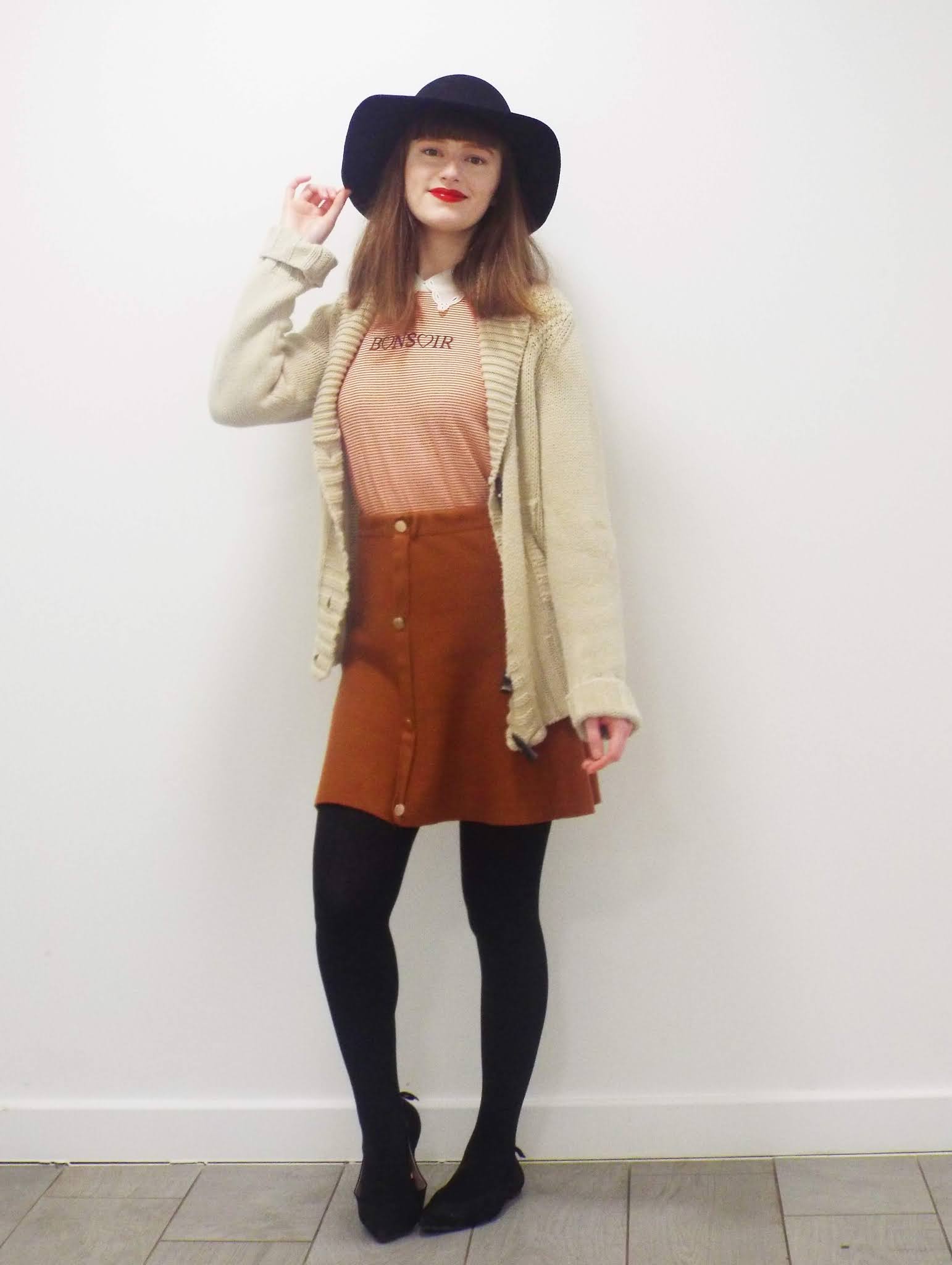 Full outfit shot of Ellie, wearing white vintage collar detailed shirt; orange and white horizontally striped T-shirt over top, that says "Bonsoir"; rust coloured mini skirt, with central gold buttons; thick black tights; black pointed toe slip on shoes; and black floppy hat, which Ellie is holding with her right hand to her head.