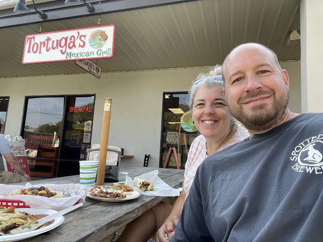 A selfie of dad and I eating lunch at Tortugas - a mexican restaurant. We are sitting outside on a picnic table. Dad and I are both sitting on the same side of the table. Dad is wearing his salty dog cafe black tshirt and I am in a pink and white striped t-shirt. We came here after our morning walk.