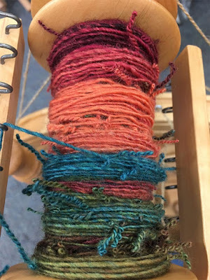 A half-full spinning wheel bobbin with bright orange, pink, green, and turquoise yarn on it, with pigtails sproinging out all over.