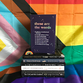 A photo of the proof of These Are the Words by Nikita Gill standing on top of a stack of other books by Nikita Gill. From top to bottom, the stack incluides Where Hope Comes From, Fierce Fairytales, Wild Embers, Great Goddesses, and The Girl and the Goddess. On top of Where Hope Comes From on the left is a rainbow pin. The stack sits on a giant Pride flag, which is also the background; the flag is pinned up, and drapes over the surface, which the stack is put on top of.