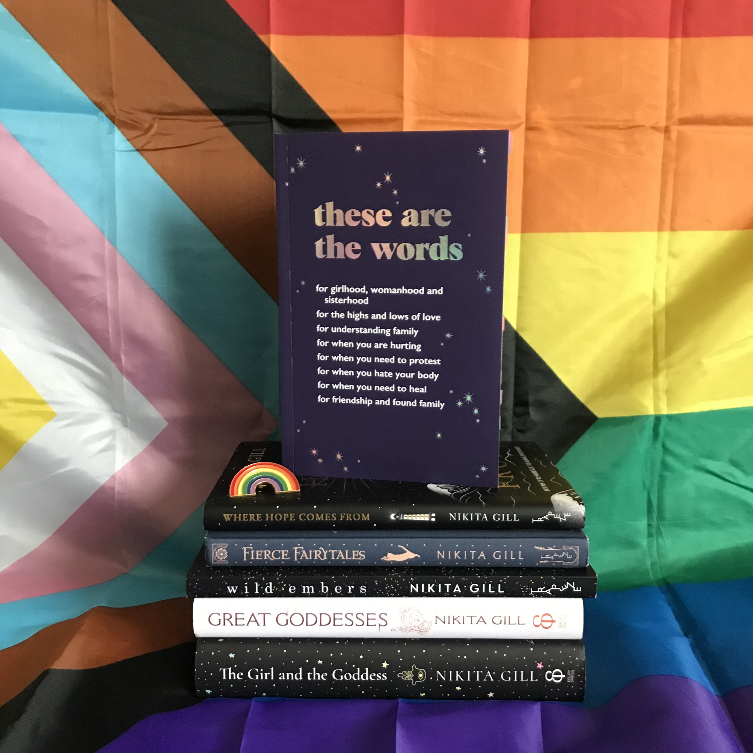 A photo of the proof of These Are the Words by Nikita Gill standing on top of a stack of other books by Nikita Gill. From top to bottom, the stack incluides Where Hope Comes From, Fierce Fairytales, Wild Embers, Great Goddesses, and The Girl and the Goddess. On top of Where Hope Comes From on the left is a rainbow pin. The stack sits on a giant Pride flag, which is also the background; the flag is pinned up, and drapes over the surface, which the stack is put on top of.