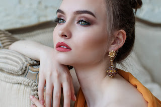 A picture of a woman with dark hair in an updo with a smoky eye and red lips sat down staring into the distance on a white background 