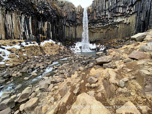 Water cascades down over hexagonal dark grey-black basalt columns into a valley covered in large boulders.