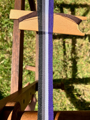 A close up of a woven band in progress on an inkle loom. The warp is striped in the colours of the ace pride flag (black, grey, white and purple). End ID.
