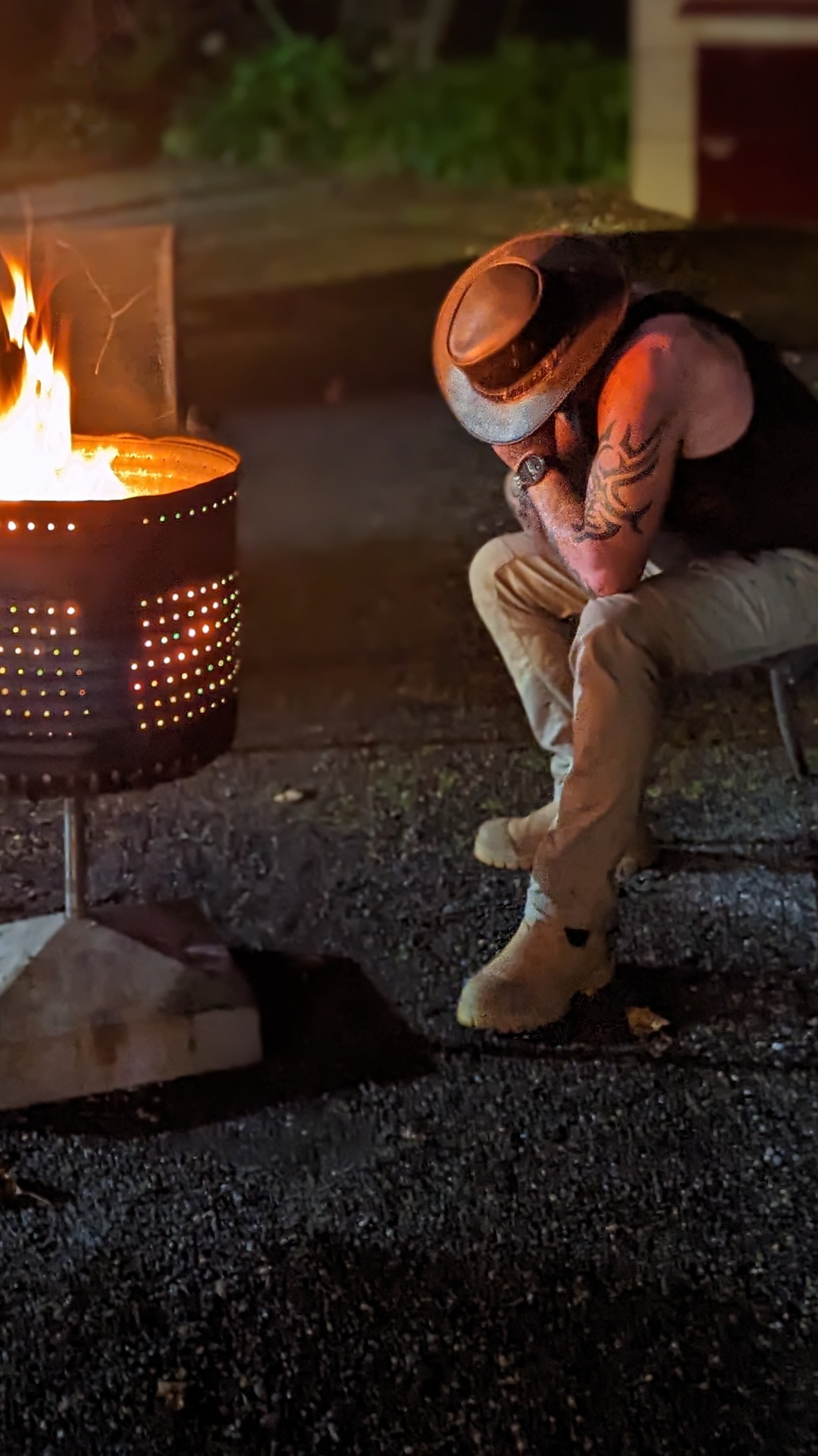 A brazier blazes and lights up a tattooed man in a leather convoy hat, black singlet, jeans, and boots as he leans forward with his head in his hands