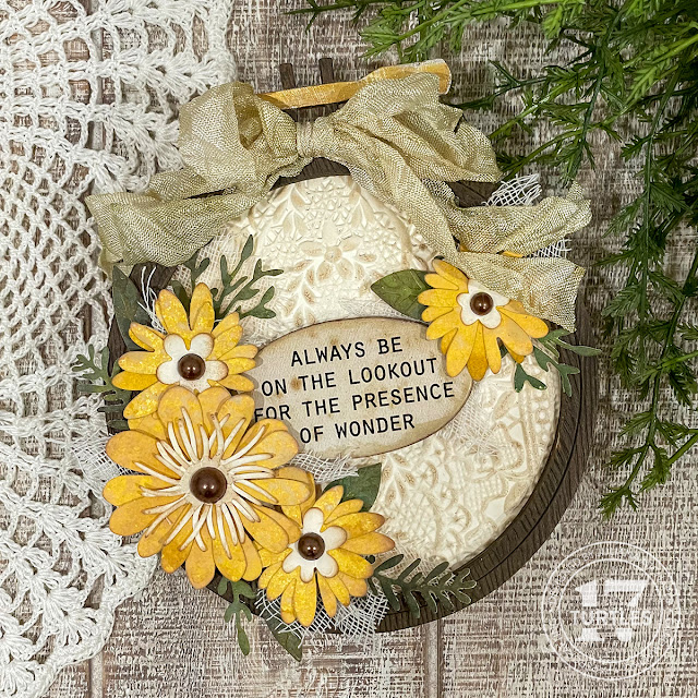 An embroidery hoop made from paper with an embossed paper lace background. Handmade yellow flowers and green leaves made from paper surround a chipboard sentiment piece that says "always be on the look out for the presence of wonder".