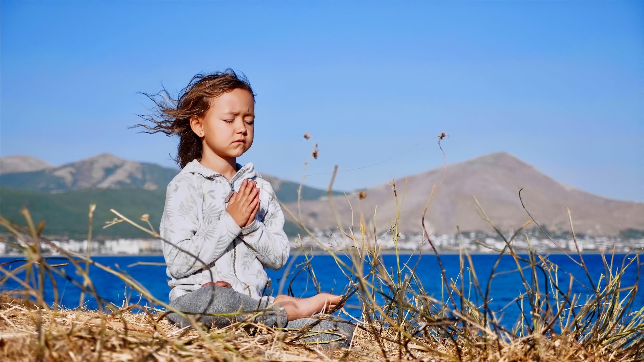 Kids Meditation - Cute little child gurl meditating alone in lotus pose at lake shore with namaskar gesture in windy