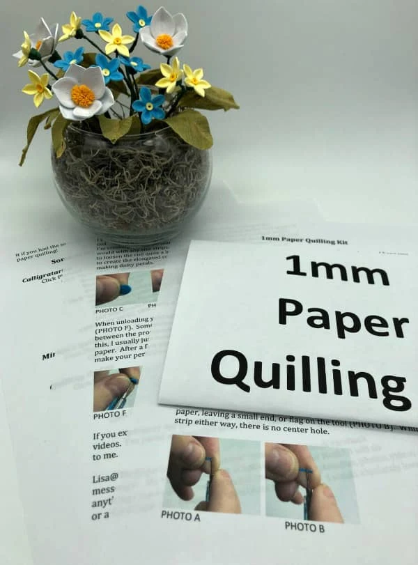 arrangement of blue, yellow, and white quilled flowers in clear vase filled with brown moss alongside quilling instructions