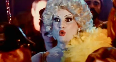 Bill in drag, with a yellow boa and a platinum blonde wig, looks surprised.