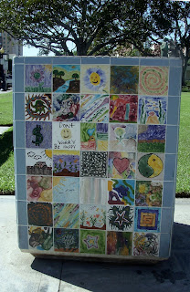 Trash can covered with hand painted tiles with different pictures and patterns.  There is a tree in the background.