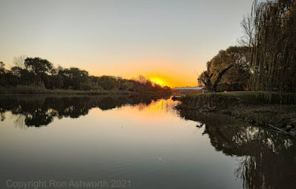 A peaceful river scene with the sun setting on the horizon by Ron Ashworth