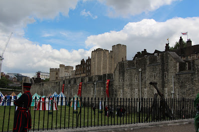 Grassy moat with striped tents, wall of the Tower of London, two of the Tower guards in black uniforms trimmed with red.