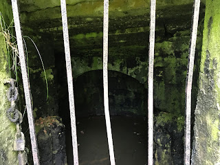 A view through the bars of the Oyly Well showing the stone structure inside and the oily waters.  Photograph by Kevin Nosferatu for the Skulferatu Project