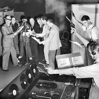 Black and white photo of six men and a woman crowding around a microphone with scripts in their hands. To the right and closer in the foreground are two men aiming smoking revolvers in the air and looking at scripts. Near the bottom of the picture is a control board with records on it.