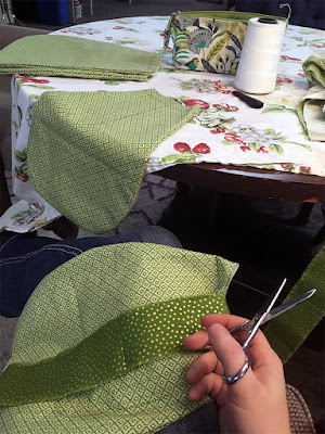 A sort of assembly line of flat-topped teardrop shapes cut from green printed fabric, laid out on a round table with a cherries and strawberries print tablecloth, a sewing pouch, and a giant spool of white thread on it. A white hand is holding small silver scissors at the very bottom of the picture.