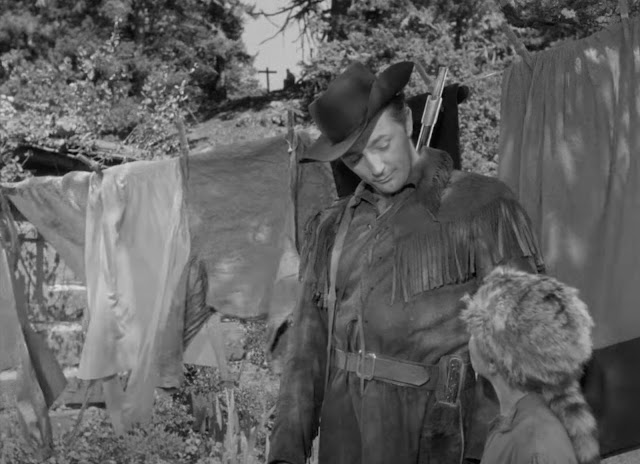 Screenshot from the 1948 movie Rachel and the Stranger showing Robert Mitchum in a cowboy hat and fringed jacket gazing down at young Gary Gray, wearing a coonskin cap. They are standing in front of laundry hanging on a wash line.