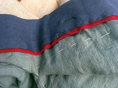 A muted teal blanket with huge, widely spaced lines of stitching in thick white thread and a layered border of deep blue with a narrow red strip tucked under the edge. A needle threaded with doubled navy thread is inserted into the blue border, halfway through stitching a large running stitch through the very edge of the blue border.