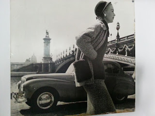 Black and white image of a woman standing in front of an old car and bridge.