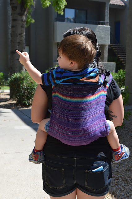 Image of fair skin toddler in a purple rainbow handwoven Onbuhimo in a back carry.  Toddler is pointing into the distance