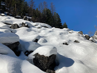 boulders on a hill covered with mounds of snow. Trees at top of hill and a patch of blue sky behind.