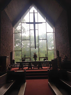 The chancel of a sanctuary in which all the lights are off. At the center is a large table, modeled after the one in the painting the Last supper. Behind that is a pentagonal floor-to-ceiling window with a large cross hanging in front of it. In the foreground, pews face towards the center aisle.