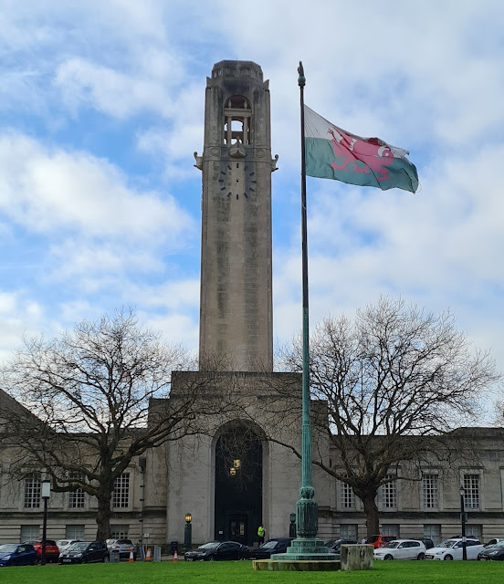 Swansea Guildhall's clock tower centre of frame with no hands on the clock itself, Welsh flag flown on the adjacent mask. Backdrop of patchy clouds with some blue sky peeping through.