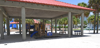 A pavilion with a red roof. The floor is made of sand. Underneath it is a playground.