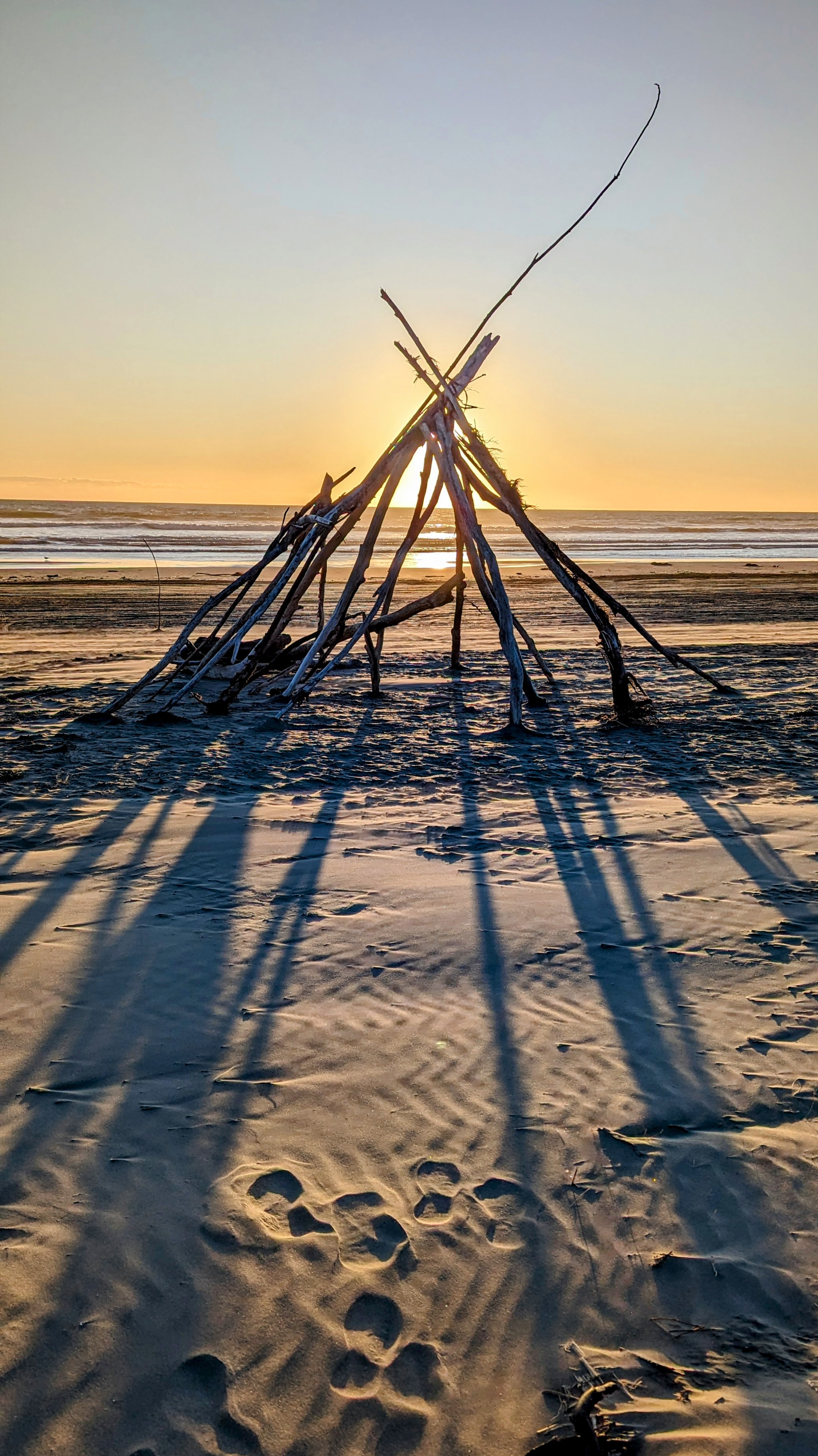 Calm beach sunset with the orange glow behind a makeshift driftwood teepee and it's elongated shadows coming towards the camera.