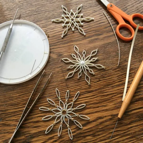 three quilled snowflakes on wood table surrounded by quilling tools