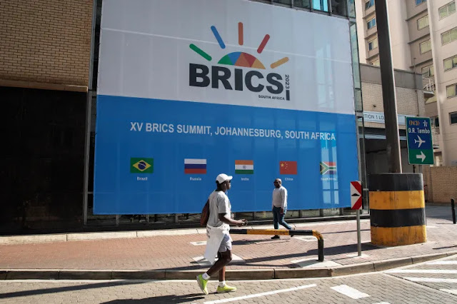 Cover Image Attribute: People walk past a banner outside the venue for the 2023 BRICS summit in Johannesburg, South Africa | Source: Gianluigi Guercia/AFP