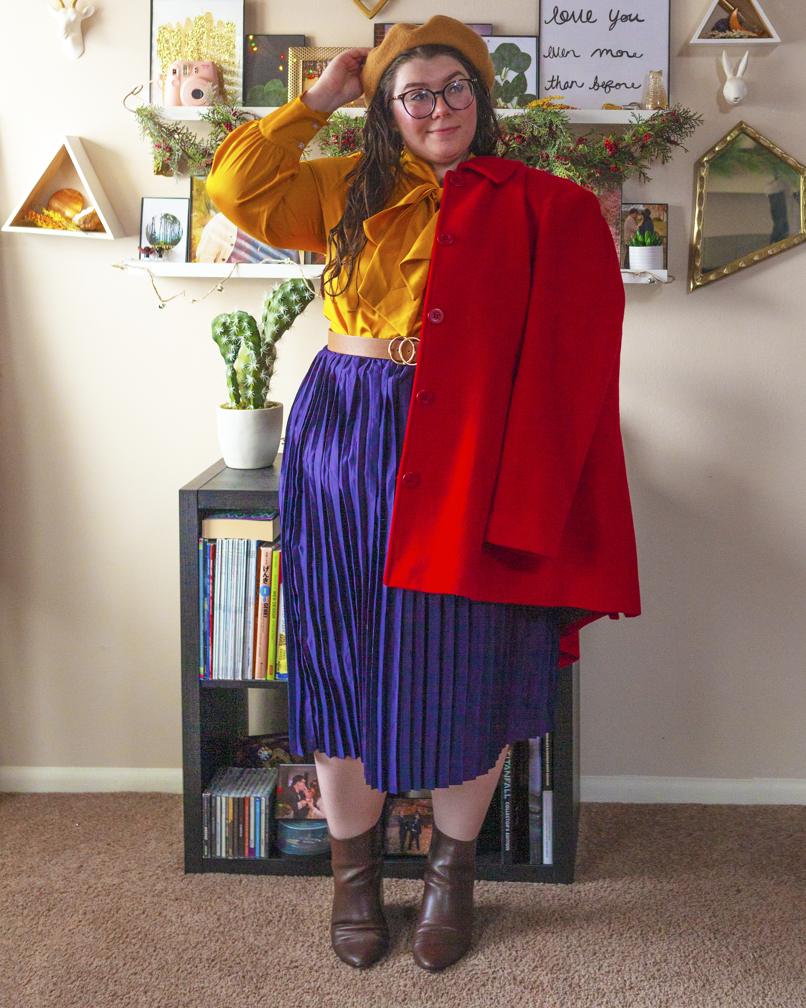 An outfit consisting of a camel brown beret, a red coat, a yellow satin long sleeve blouse tucked into a navy blue pleated midi skirt and sheer black tights and brown boots.