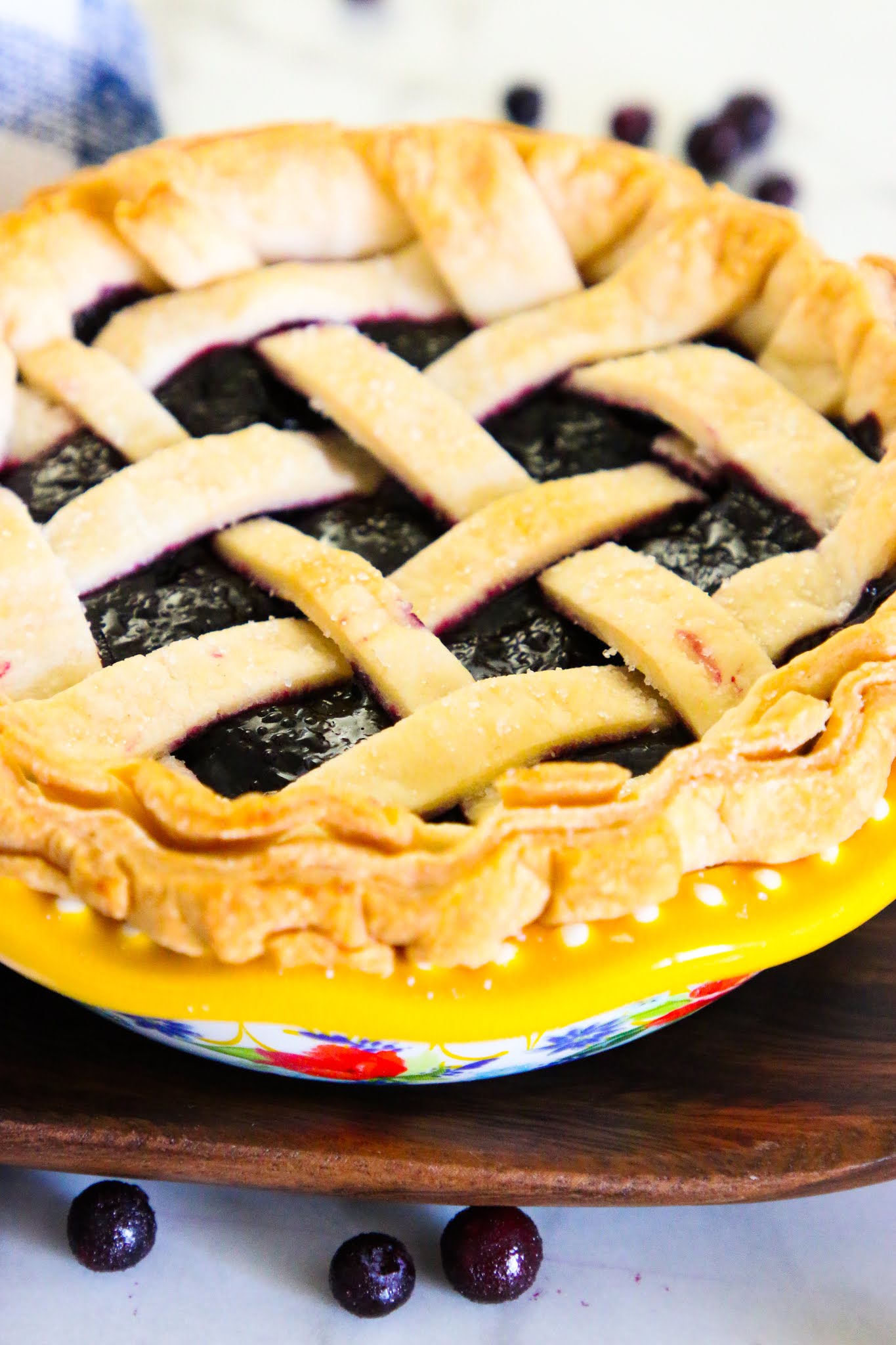 Blueberry pie in a blue pie plate with a blue dish towel in the background and blueberries scattered on the table.