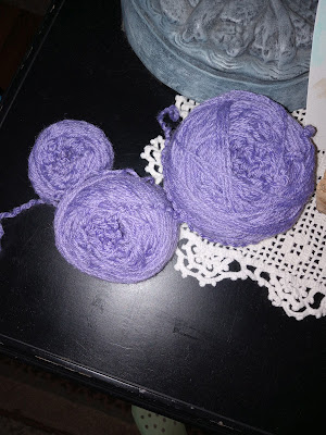 Three balls of lilac purple yarn, each larger than the next, rest on a black wooden console with a white crocheted lace doily in the background.