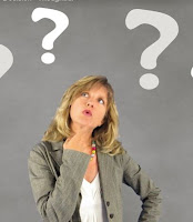 A photo of a woman with blonde hair. She is wearing a grey jacket and white top. Above her head are question marks.  She looks quizzical.