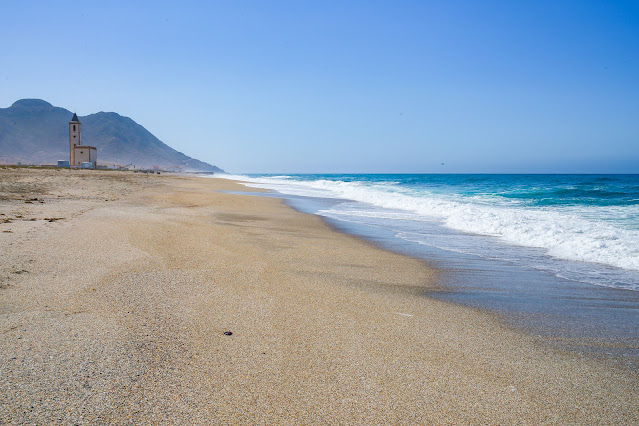 Playa de arena dorada y mar de azules aguas con una iglesia y montañas al fondo