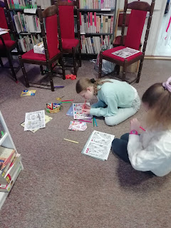 Biblioteka. Na wykładzinie dywanowej siedzą dwie dziewczyny. Kolorują litery z baneru. W tle regały biblioteczne.