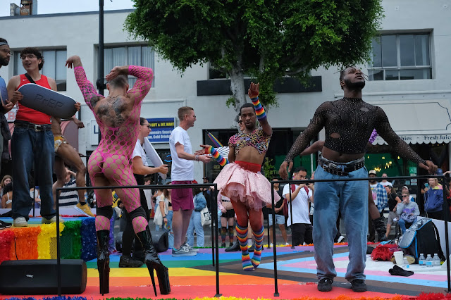 A pride parade float with multiple men dancing enthusiastically.  One is dressed in pink netting, another wears a leopard print crop top and puffy pink skirt, another is in jeans and a black shirt.