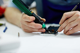A volunteer at a Repair Cafe fixes a broken cellphone, demonstrating the hands-on approach to reducing electronic waste and promoting sustainability. The image highlights the community effort to extend the life of electronic devices, a key aspect of Repair Cafes in fostering a sustainable and resourceful community.