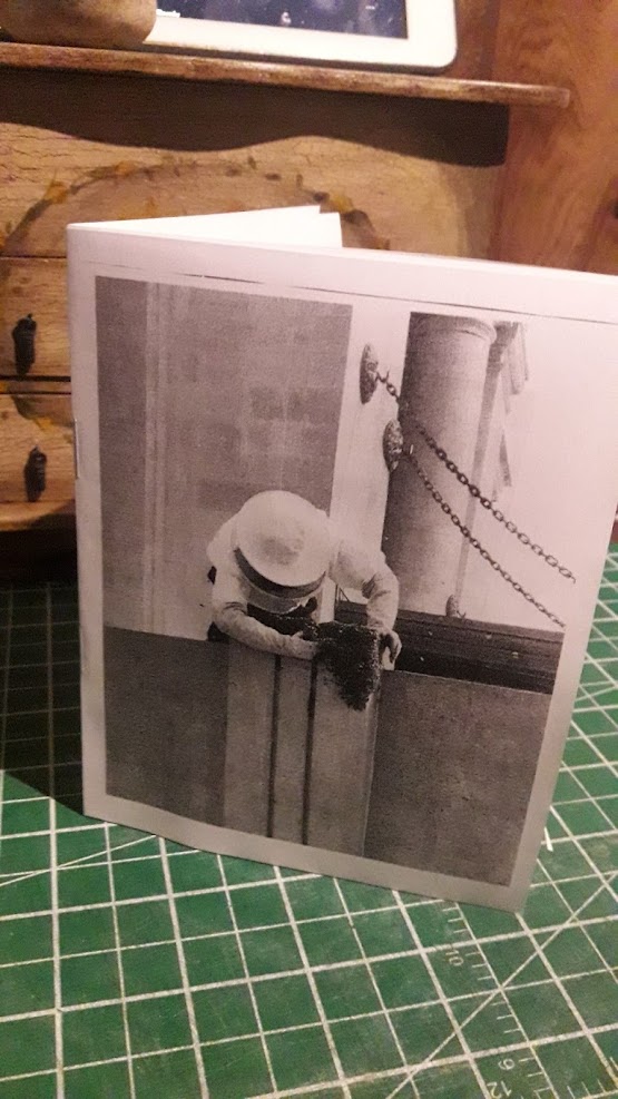 photo of a quarter-letter size zine standing on a cutting board. the cover is a black & white photo of a beekeeper removing a hive from a stone building