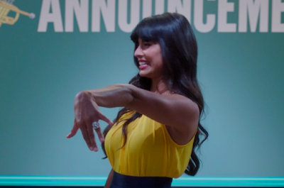 Tahani showing off an enormous engagement ring. She's wearing a yellow sleeveless top and is in front of the superboard, which says "ANNOUNCEME" (the NT got cut off)