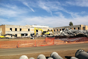 Front of building pictured with 'B' wing demolished, windows removed, rubble, and heavy equipment in the foreground.